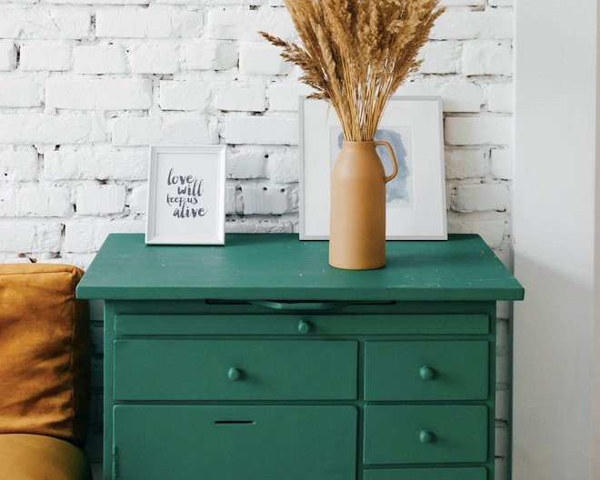 A book, brown flower vase with wheat inside, and a picture frame on top of a pale green drawer